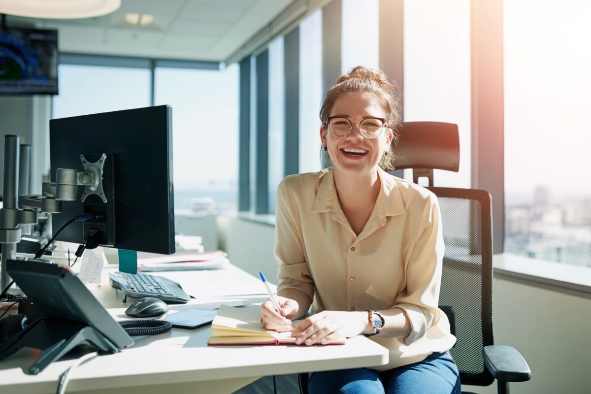 Woman, smiling, sitting at a computer desk, writing notes on a notebook
