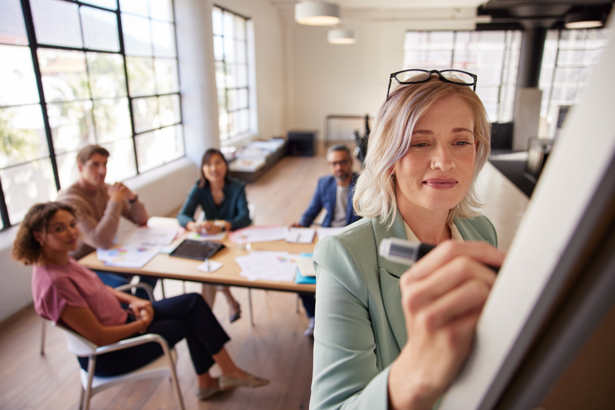 Woman writing notes on a white board, whilst others sit at desk behind her, business meeting setting.