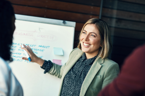 Woman smiling, in a meeting setting, writing notes on a white board and talking to a few other people
