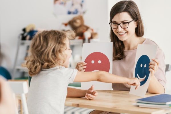 Woman smiling, sat at a desk with a small child. She's showing the child two pieces of paper, one with a sad emoticon and one with a happy emoticon. The child points at the happy emoticon.