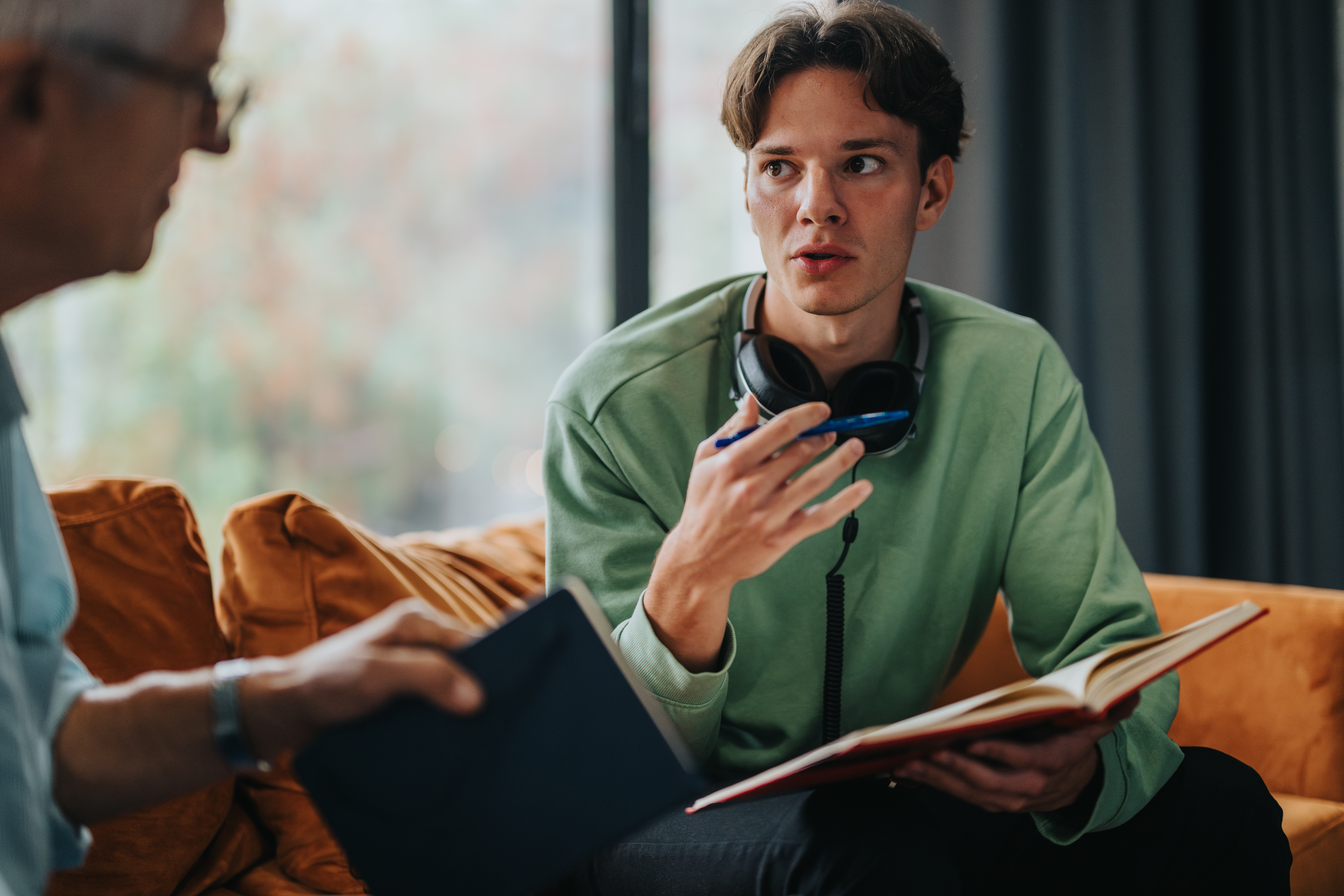 A young man with headphones around his neck converses with a mentor while holding a book.
