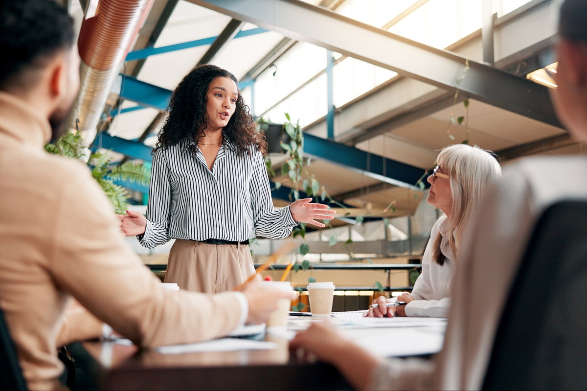 Professional meeting, with a woman stood up talking to a few people that sit around a desk listening