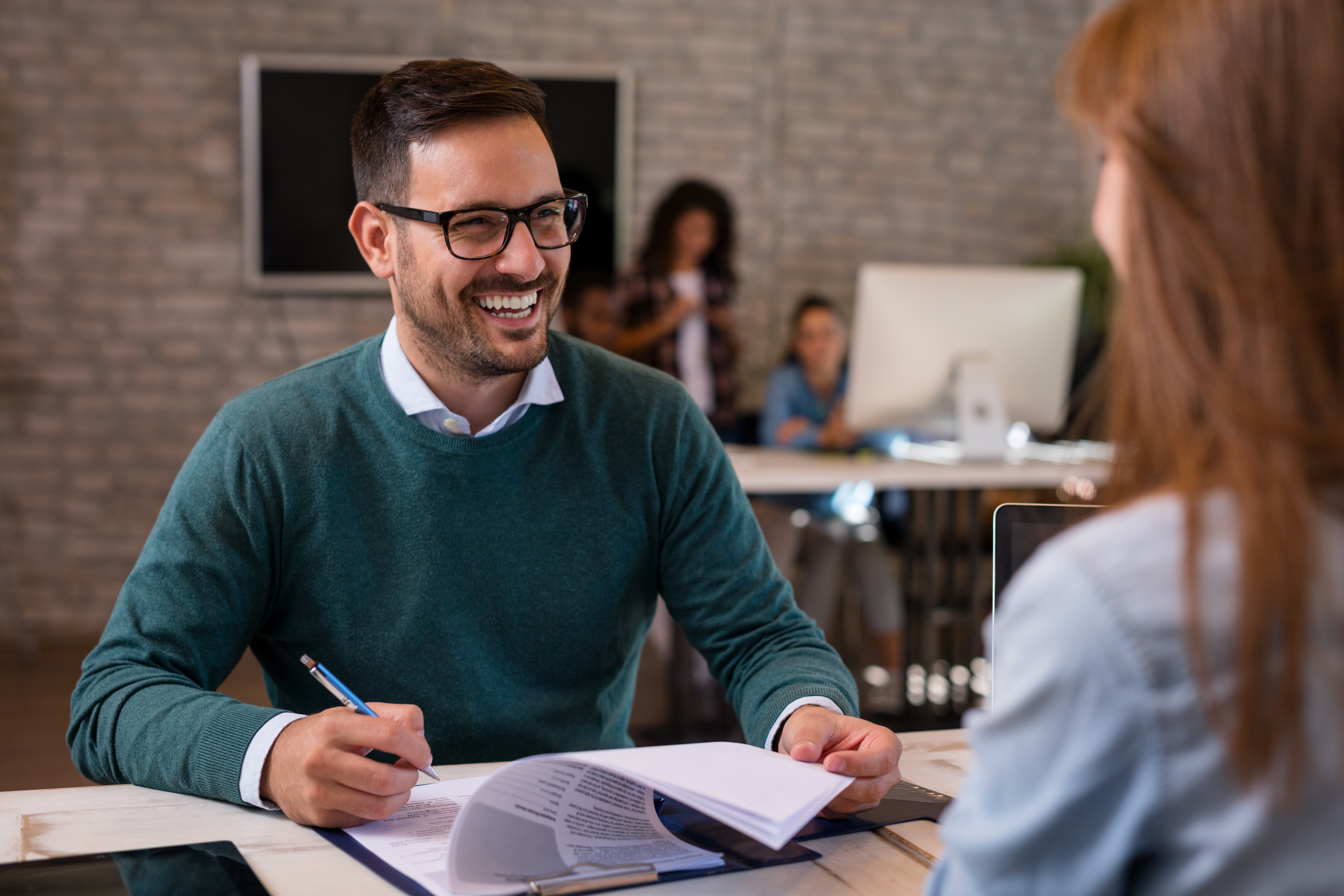 Man smiling, sat across from a woman, he's holding a clipboard and a pen