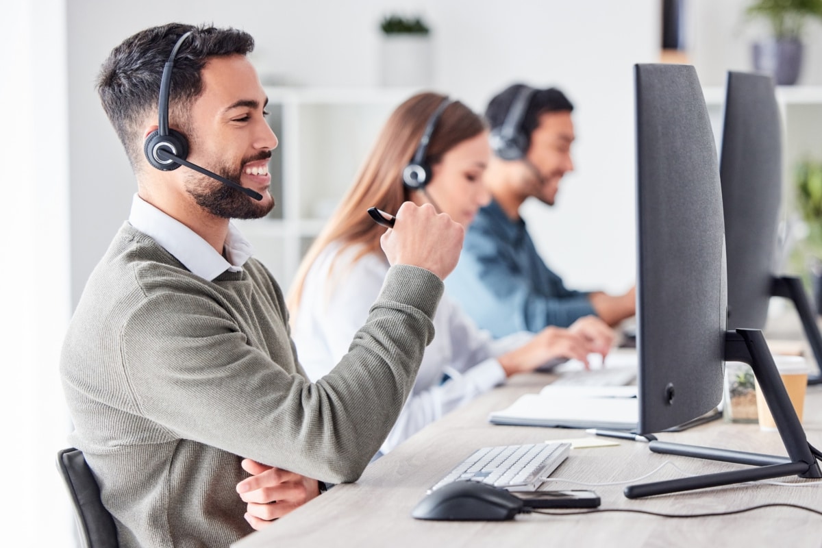 Three people working in call centre. They all have headsets on and are sat in front of a desktop computer