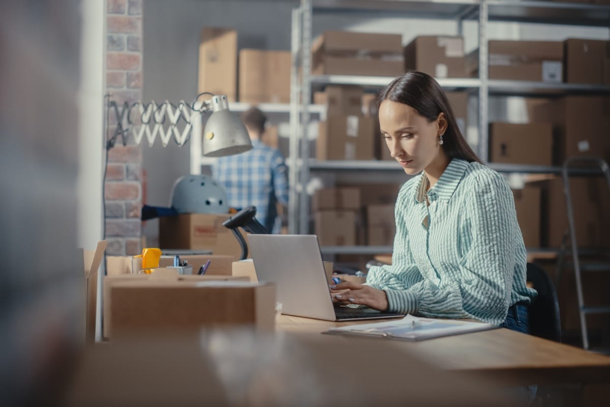 Online store inventory manager using laptop computer, preparing a small parcel for postage.