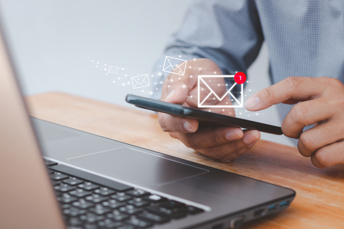 Close up of someone using a laptop and mobile phone. Icon of email is overlayed.