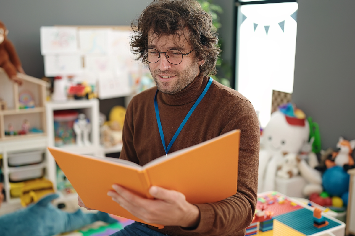 Male early years teacher reading book standing in nursery