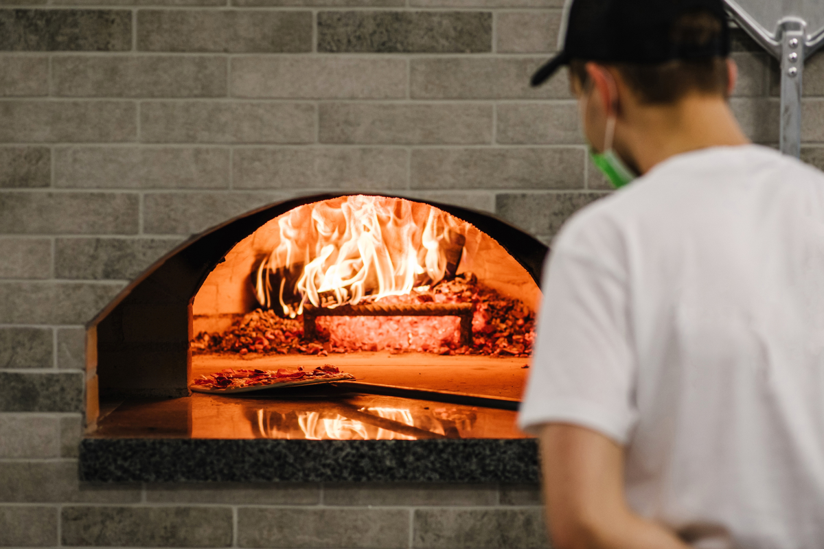 A chef apprentice puts a pizza on a shovel in the wood fire pizza oven.